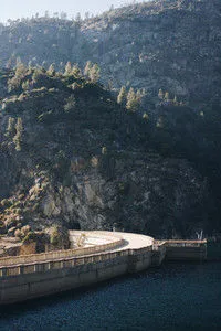 Curved concrete bridge over dark water with steep rocky hillside covered in scattered trees in soft daylight