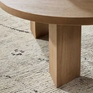 Close-up showing round light-brushed parawood tabletop with thick triangular pedestal leg on textured rug