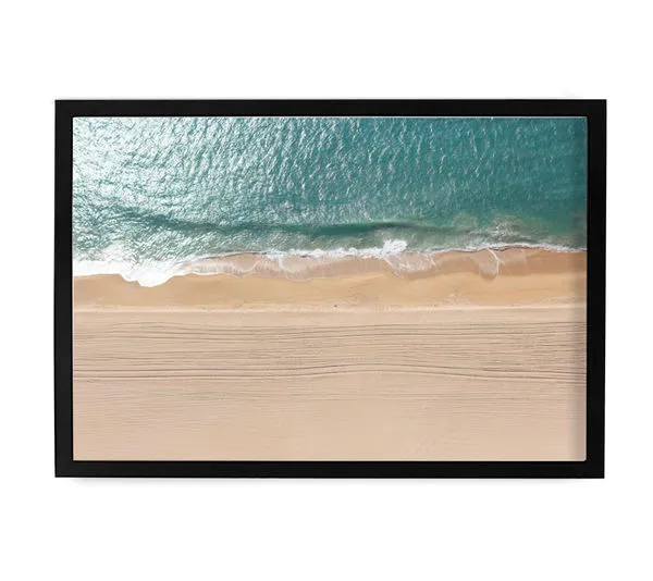 Aerial ocean photography showing blue waves meeting a sandy beach, framed in a sleek black frame