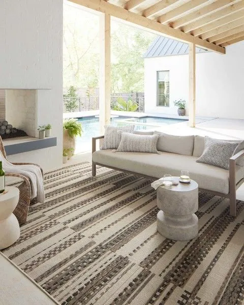 Rug laid out under neutral-toned outdoor sofa on covered patio near swimming pool and fireplace