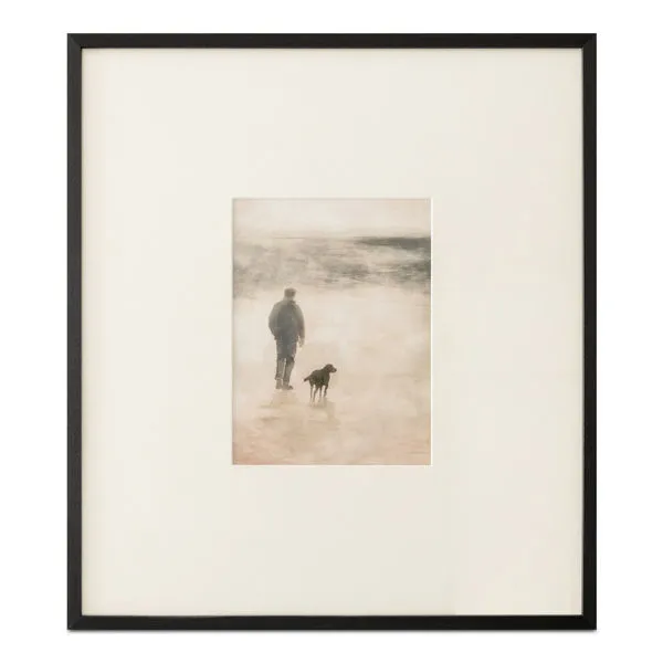 Framed print of man and dog walking on misty shore in earthy tones with black wood frame and textured matte