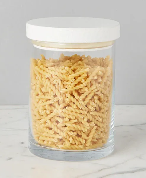 Single clear glass canister with smooth white wood top containing spiral pasta on marble surface