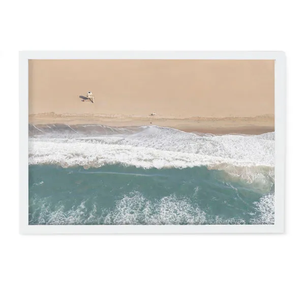 Aerial beach photography featuring a sandy shore, foamy waves, and a small lifeguard tower casting a shadow