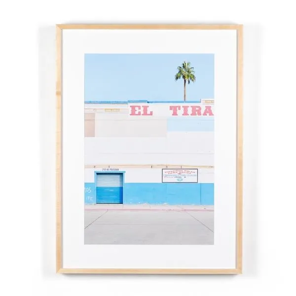 Framed photograph of a building facade with blue and white walls, pink signage, and a single palm tree