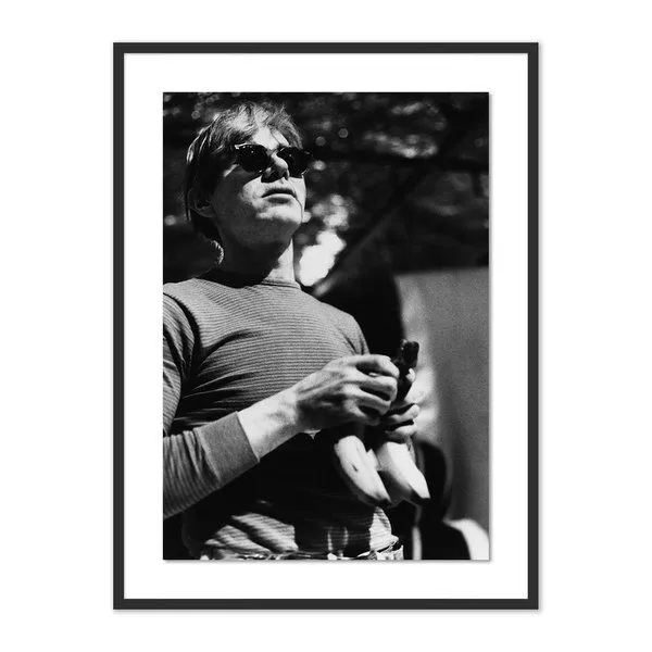Black and white photo of Andy Warhol wearing sunglasses and holding bananas, framed in black with white matting