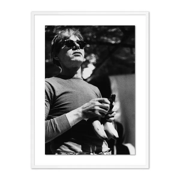 Black and white photo of Andy Warhol wearing sunglasses holding bananas, framed with white mat and frame