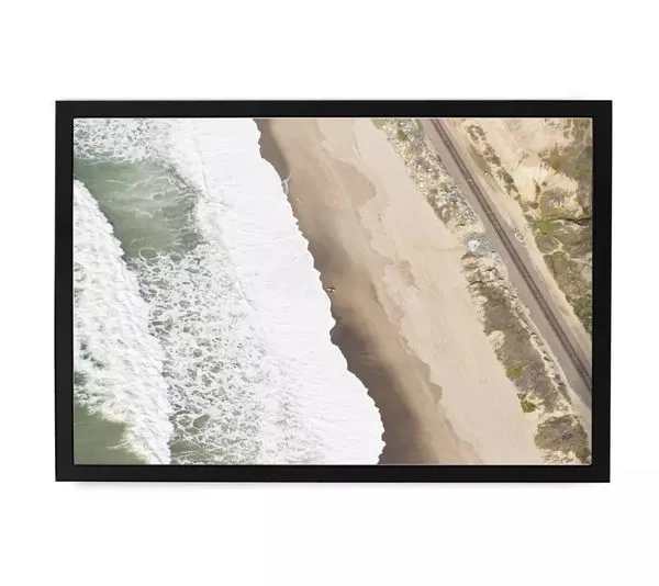 Aerial seaside photograph showing a lone surfer waiting to paddle out, waves crashing on sandy beach near railway track