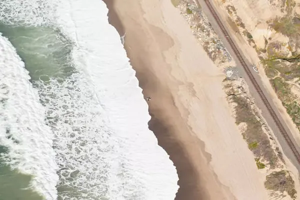 Aerial view of a lone surfer waiting on a sandy beach with rolling ocean waves and nearby railway tracks at Riviera Beach ...