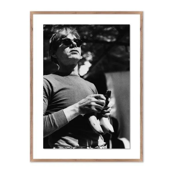 Black and white photograph of Andy Warhol wearing sunglasses and holding bananas, framed with a wooden border